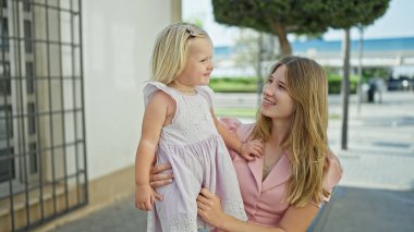Neşeli beyaz anne ve kızı sıcak, sevgi dolu bir kucaklaşmayı kucaklıyor, bir bankta oturuyor, birlikte gülüyor ve gülümsüyor, neşe ve mutlulukla dolu, güneşli bir sokakta..