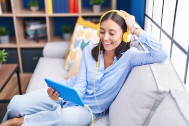 Young beautiful hispanic woman watching video on touchpad sitting on sofa at home