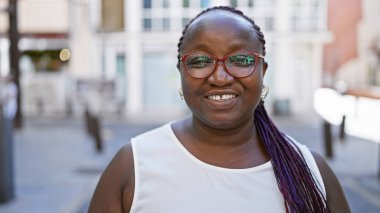 Kendine güvenen Afro-Amerikalı kadın, şehir sokaklarında duruyor, neşe ve pozitiflik saçıyor, bulaşıcı gülüşü mutluluk saçıyor. Saçları zıplıyor, gözlükleri parıldıyor, hayatı gerçekten seviyor..