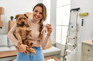 Young beautiful hispanic woman hugging dog holding key at new home