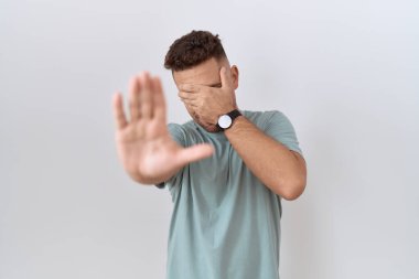 Hispanic man with beard standing over white background covering eyes with hands and doing stop gesture with sad and fear expression. embarrassed and negative concept. 