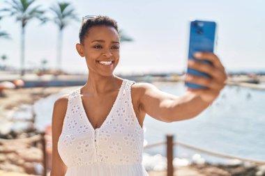 Afro-Amerikalı kadın, deniz kenarındaki akıllı telefonun yanında kendinden emin bir şekilde selfie çekiyor.