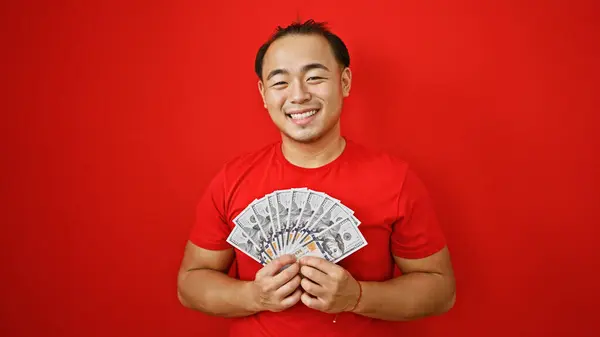 Confident young chinese man, gleaming with happiness, casually holding a heap of us dollars, smiling boldly over an isolated red background.