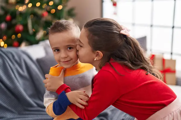 Brother and sister kissing and hugging each other standing by christmas tree at home