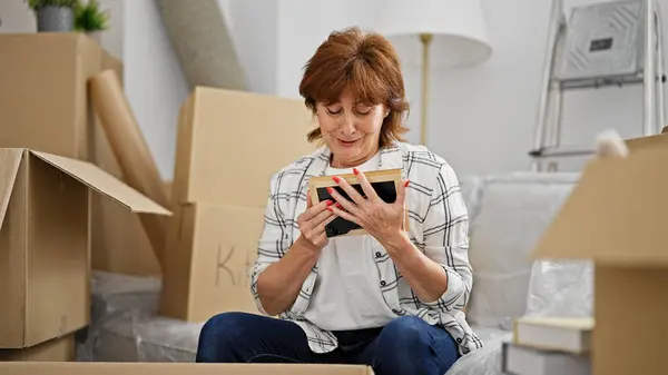 Middle age woman unpacking cardboard box looking photo at new home