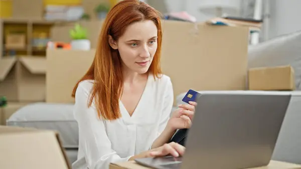 Young redhead woman shopping with laptop and credit card sitting on floor at new home