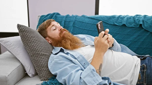 英俊的 留着胡子的红头发的男人 完全放松而严肃 专心致志地使用自己的手机 躺在家里的沙发上 在舒适的客厅里发短信 迷失在网络技术的世界里 — 图库照片