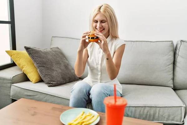 年轻的金发女人坐在家里的沙发上吃汉堡包 — 图库照片