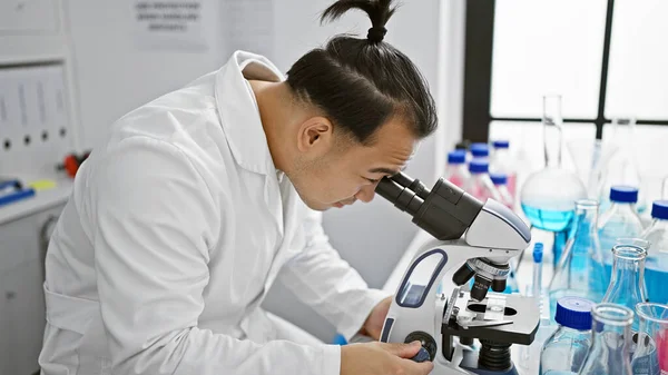 Serious young chinese man, a professional scientist, concentrating at a microscope in a bustling lab - a marriage of medicine and technology