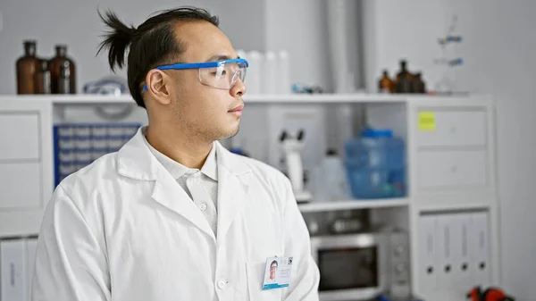 Serious young chinese man, a lab scientist in safety glasses, deep in concentration on medical research at a bustling, professional chemistry lab.