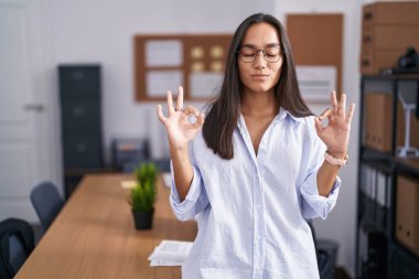 Ofisteki genç İspanyol kadın rahatlamış ve gözleri kapalı gülümsüyor parmaklarıyla meditasyon hareketi yapıyor. yoga konsepti. 
