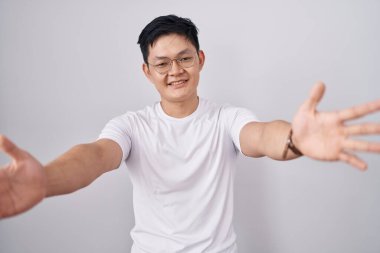 Beyaz arka planda duran Asyalı genç adam kameraya bakıp sarılmak için kollarını açmış gülümsüyor. Mutluluğu kucaklayan neşeli bir ifade.. 