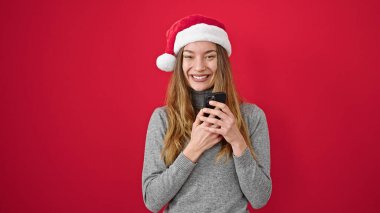 Kafkasyalı genç bir kadın noel şapkası takıyor ve izole edilmiş kırmızı arka plan üzerinde akıllı telefon kullanıyor.