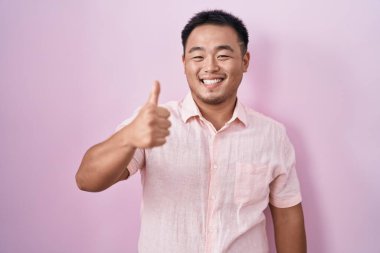 Pembe arka planda duran Çinli genç adam elleriyle mutlu bir jest yapıyor. Kameraya bakıp başarıyı gösteren ifade. 