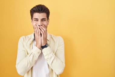 Young hispanic man standing over yellow background laughing and embarrassed giggle covering mouth with hands, gossip and scandal concept 