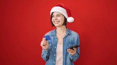 Akıllı telefon ve kredi kartı kullanan beyaz bir kadın izole edilmiş kırmızı arka planda noel şapkası takıyor.