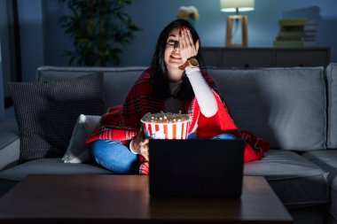 İspanyol kadın kanepede film seyrederek patlamış mısır yiyor bir gözünü eliyle kapatıyor, yüzünde kendine güvenen bir gülümseme ve sürpriz bir duygu var.. 