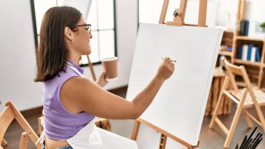 Young beautiful hispanic woman artist drinking coffee drawing at art studio