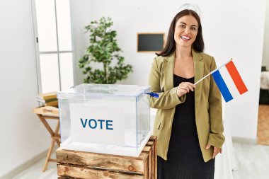 Young beautiful hispanic woman smiling confident holding netherlands flag standing at electoral college