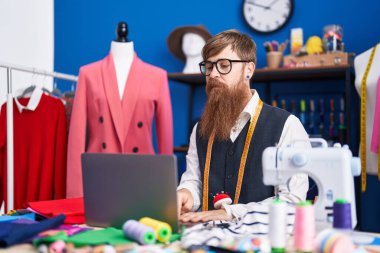 Young redhead man tailor using laptop at clothing factory