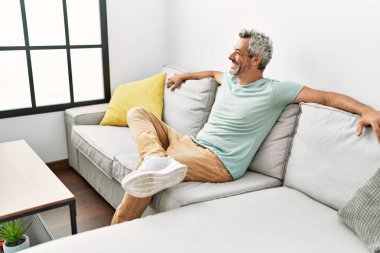 Orta yaşlı, kır saçlı, gülümseyen bir adam. Evdeki kanepede oturuyor.