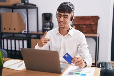 Bilgisayarda çalışan genç İspanyol bir adam. Elinde büyük bir gülümseme olan bir bilgisayar kartı var. Kameraya el ile işaret ediyor.. 