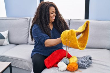 Afro-Amerikalı kadın kanepeye oturmuş evde el yapımı giysiler dikiyor.