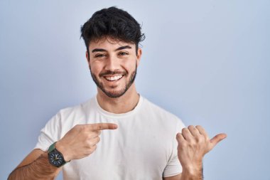 Beyaz arka planda duran sakallı İspanyol bir adam. Arka tarafı işaret ediyor. El ve baş parmakları havada, kendinden emin gülümsüyor. 
