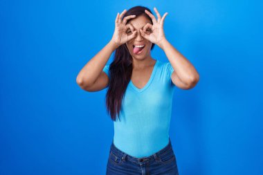 Mavi arka planda duran genç İspanyol kadın dürbün gibi jest yapıyor, gözleri parmaklarından bakıyor. çılgın ifade. 