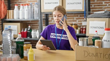 Young blonde woman volunteer talking on smartphone using touchpad at charity center clipart