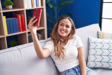 Young beautiful hispanic woman make selfie by smartphone sitting on sofa at home