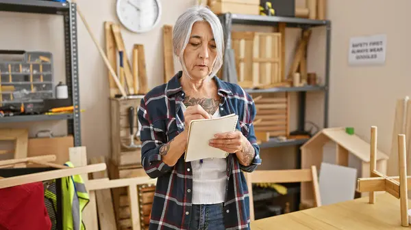 严肃而成熟的灰头发女木匠在她活泼的木工工作室里做详细的笔记 — 图库照片