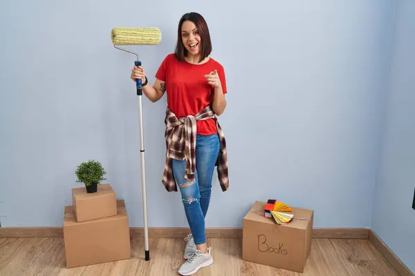 Young hispanic woman painting home walls with paint roller pointing fingers to camera with happy and funny face. good energy and vibes.