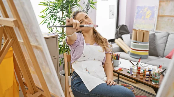 一个穿着围裙的年轻漂亮的惊慌失措的女画家 看着手中的画笔沉思不已 想着如何在舒适的画室里创作她的下一部画布杰作 — 图库照片