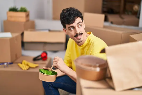 Young caucasian man eating salad sitting on sofa at new home