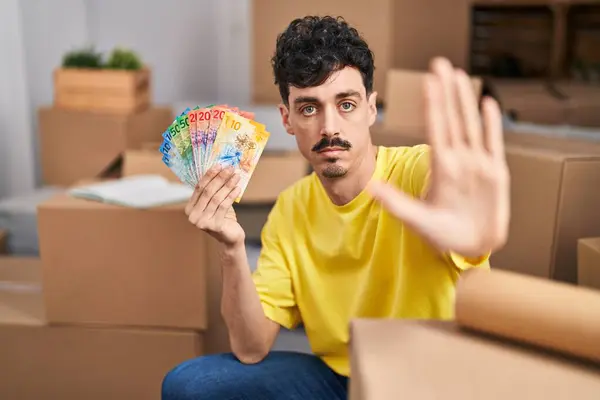 Hispanic man at new new home holding swiss francs banknotes with open hand doing stop sign with serious and confident expression, defense gesture