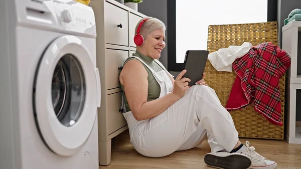 Middle age grey-haired woman watching video on touchpad waiting for washing machine at laundry room