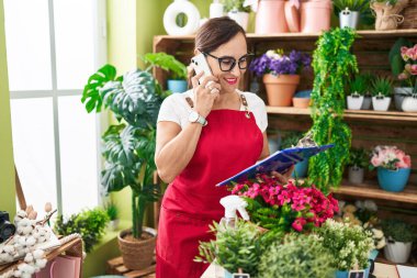 Young beautiful hispanic woman florist talking on smartphone reading clipboard at flower shop