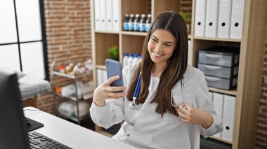 Genç ve güzel İspanyol kadın doktor klinikte gülümseyen akıllı telefonuyla selfie çekiyor.