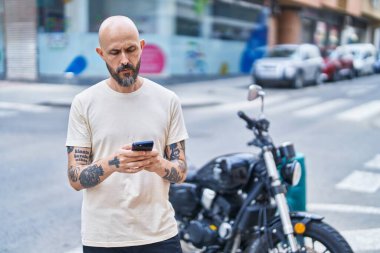 Genç kel adam akıllı telefon kullanıyor ve sokakta ciddi bir yüz ifadesi var.