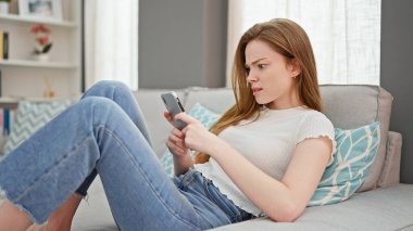 Akıllı telefon kullanan genç sarışın kadın evde üzgün görünüyor.