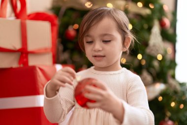 Evde Noel ağacının yanında duran çok tatlı bir kız elinde balo süsü tutuyor.