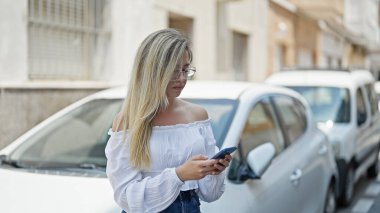 Sokakta arabaya yaslanmış akıllı telefon kullanan genç sarışın kadın.