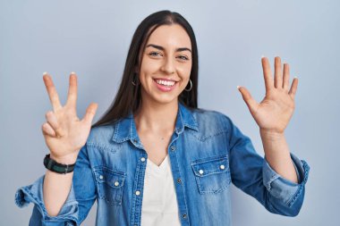 Hispanic woman standing over blue background showing and pointing up with fingers number eight while smiling confident and happy. 