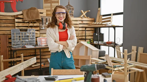 Confident young woman, a professional carpenter, standing relaxed in her woodworking studio, arms crossed, wearing security glasses with a smile