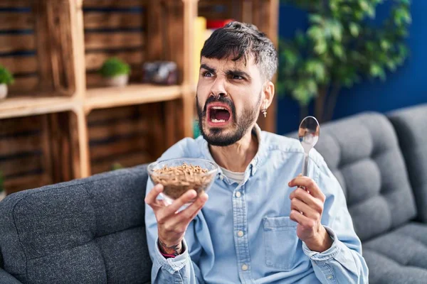 Young hispanic man with beard eating healthy whole grain cereals angry and mad screaming frustrated and furious, shouting with anger. rage and aggressive concept.
