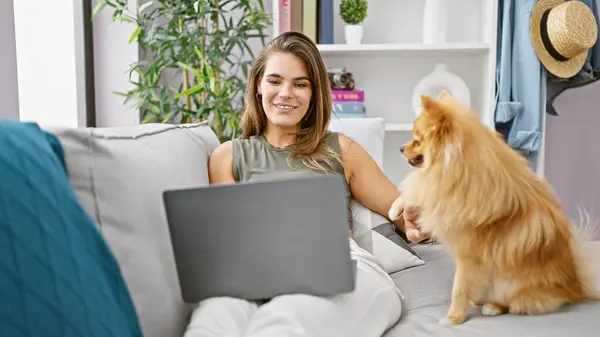 快乐而又惊慌的年轻女人坐在沙发上 和她的爱玩的狗一起使用笔记本电脑 在家里的客厅里享受在线技术的舒适 — 图库照片