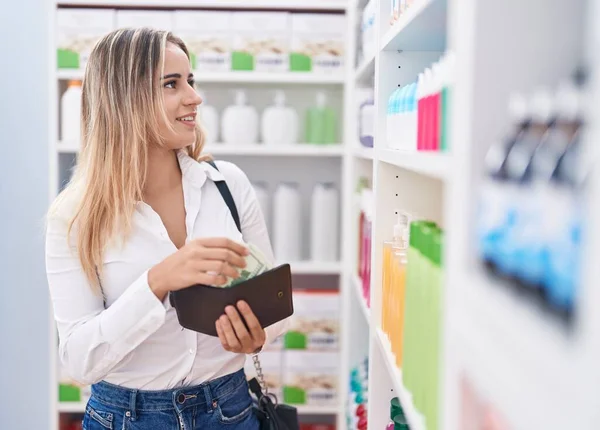 Young blonde woman customer smiling confident holding dollars of wallet at pharmacy