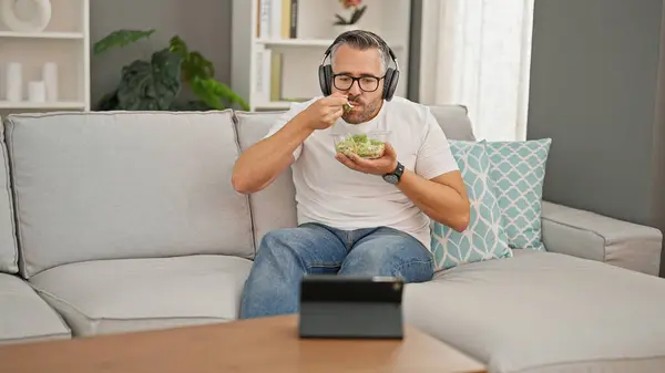 Grey-haired man eating salad watching video on touchpad at home