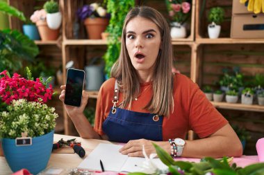 Çiçekçide çalışan İspanyol asıllı genç bir kadın akıllı telefon ekranında korkmuş ve ağzı açık bir şekilde şaşırmış, inançsız yüz ifadesi sergiliyor. 